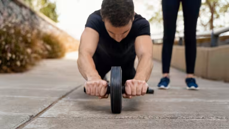 Man in black t-shirt using ab wheel on pavement, focused downward, with person's lower legs visible behind him.