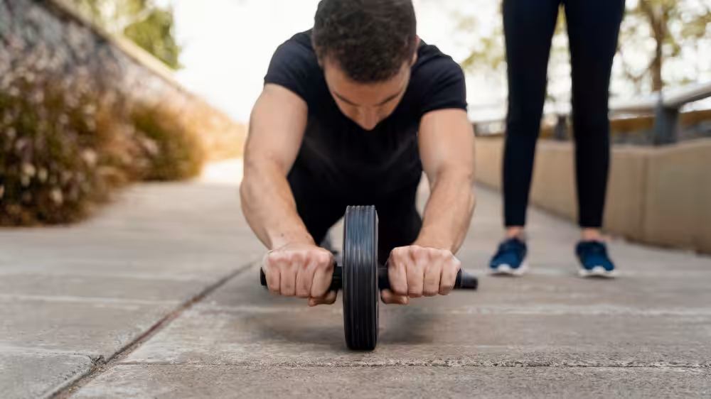 Man in black t-shirt using ab wheel on pavement, focused downward, with person's lower legs visible behind him.