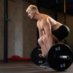 Muscular blonde man in black shorts performing deadlift with 20 kg weight plates in industrial gym with concrete floors.
