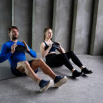 Man in blue shirt and woman in black workout clothes doing sit-ups with weights on gray mats in industrial gym space.