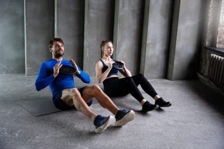 Man in blue shirt and woman in black workout clothes doing sit-ups with weights on gray mats in industrial gym space.