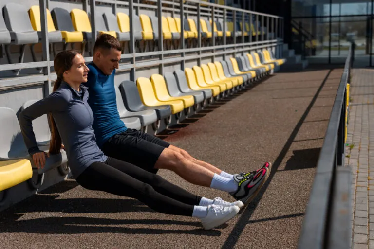 A man and woman in athletic wear do triceps dips on stadium seats outdoors.