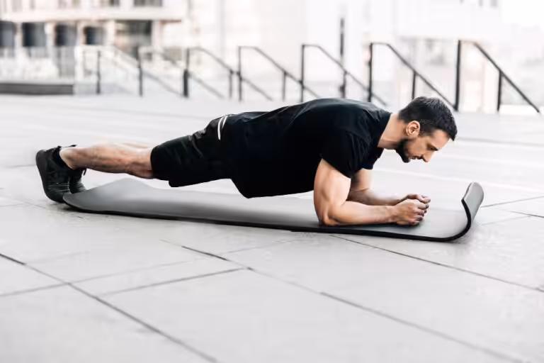 Man in black athletic wear doing plank exercise on gray yoga mat outdoors on concrete with white stairs behind.