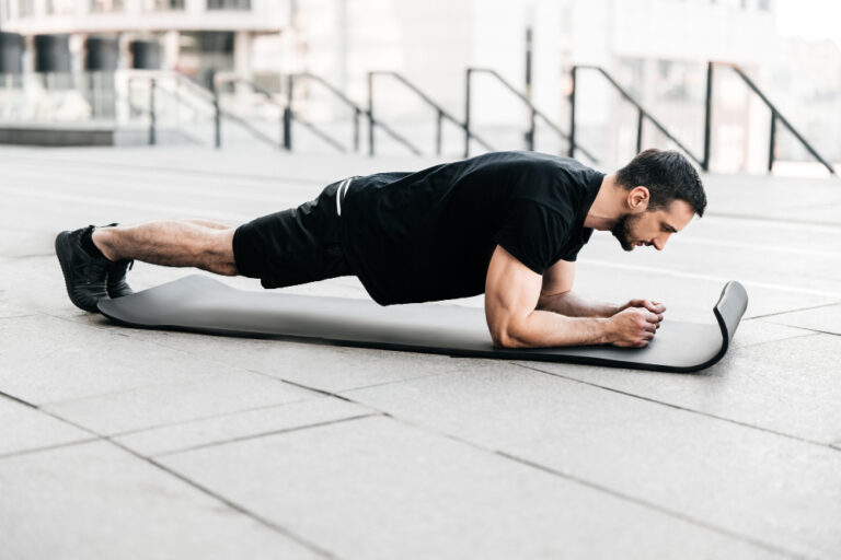 Man in black athletic wear doing plank exercise on gray yoga mat outdoors on concrete with white stairs behind.