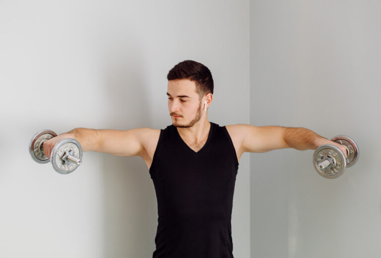 Man in black tank top performs lateral raises with dumbbells, arms extended horizontally against white wall.
