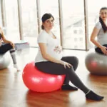 Three pregnant women in workout clothes sitting on exercise balls in bright studio with large windows and wooden floor.