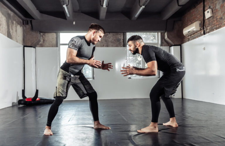 Two men in athletic wear sparring barefoot on black mats in industrial gym with brick walls and exposed ceiling beams.
