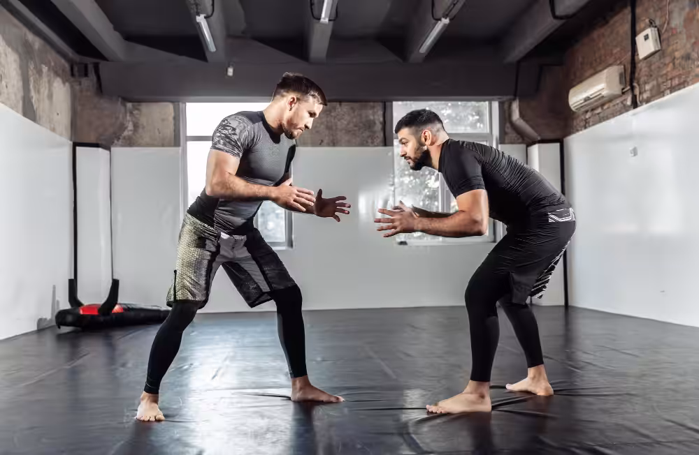 Two men in athletic wear sparring barefoot on black mats in industrial gym with brick walls and exposed ceiling beams.