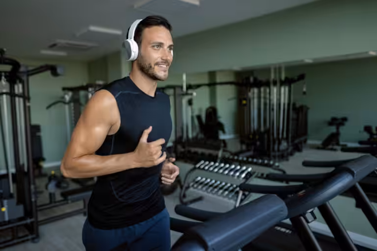 Smiling bearded man in black tank top and white headphones running on treadmill in gym with green walls.