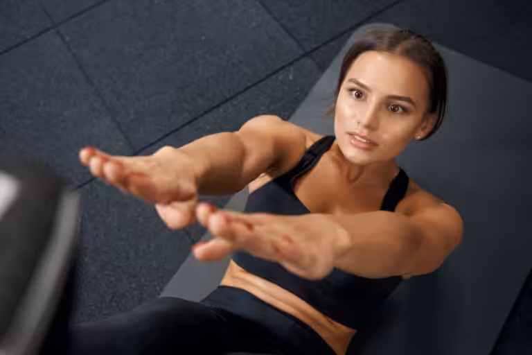 Overhead view of woman in black sports bra doing sit-up exercise on gray gym mat reaching arms forward toward camera.
