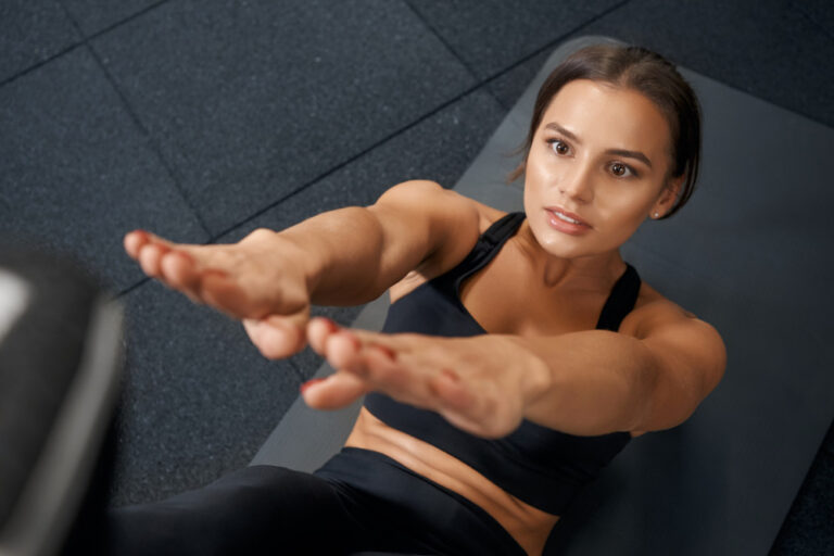 Overhead view of woman in black sports bra doing sit-up exercise on gray gym mat reaching arms forward toward camera.