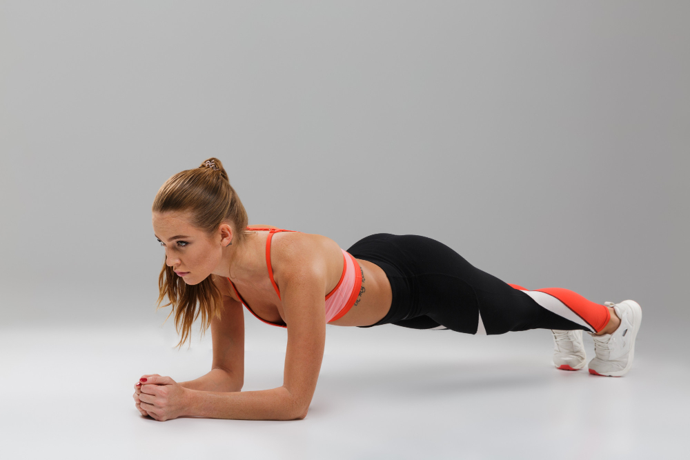 A woman with a brown pony tail in black leggings and a two-color tank top does a plank against a white background.