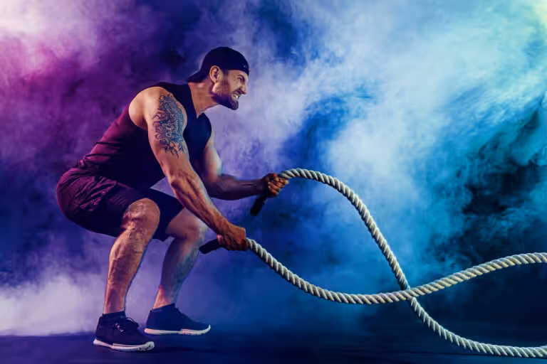 Tattooed athlete in black tank performs battle rope exercise against vibrant purple and turquoise smoke background.