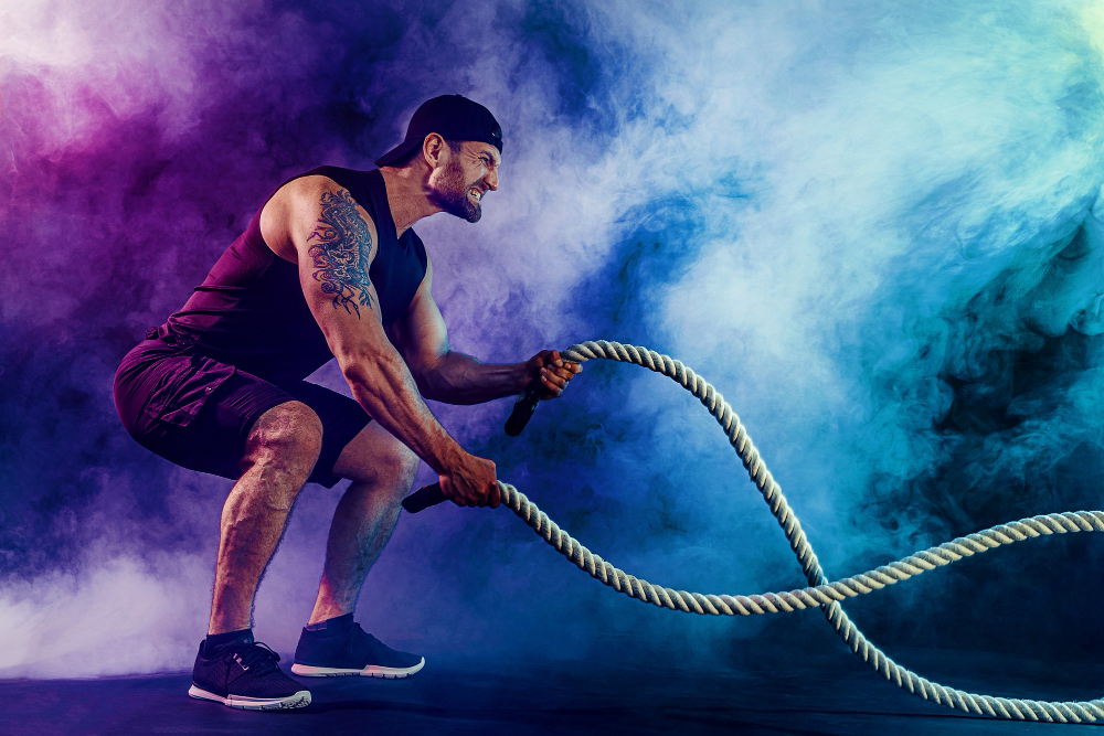Tattooed athlete in black tank performs battle rope exercise against vibrant purple and turquoise smoke background.