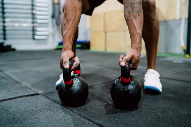 Man with tattooed arms holding two black kettlebells with red handles in gym.