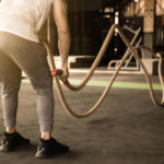 Person in white tank top and gray leggings using battle ropes in outdoor gym area.