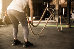 Person in white tank top and gray leggings using battle ropes in outdoor gym area.