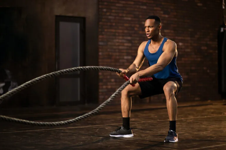 Man in blue tank top with red-handled battle rope in athletic stance inside brick-walled gym.