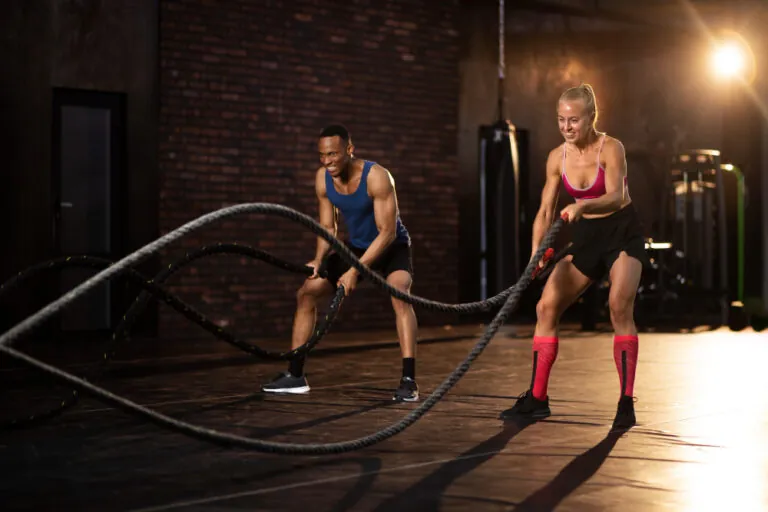 Man in blue tank top and woman in pink sports bra using battle ropes in industrial gym with brick walls and overhead lighting.