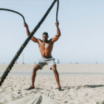 Shirtless man in gray shorts using battle ropes on sandy beach with clear sky.