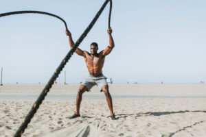 Shirtless man in gray shorts using battle ropes on sandy beach with clear sky.