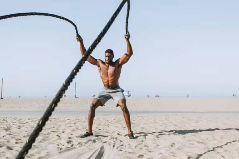 Shirtless man in gray shorts using battle ropes on sandy beach with clear sky.