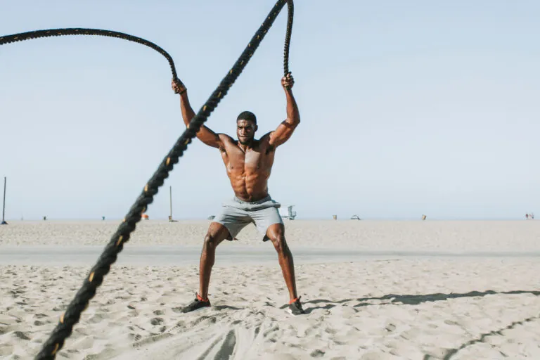 Shirtless man in gray shorts using battle ropes on sandy beach with clear sky.