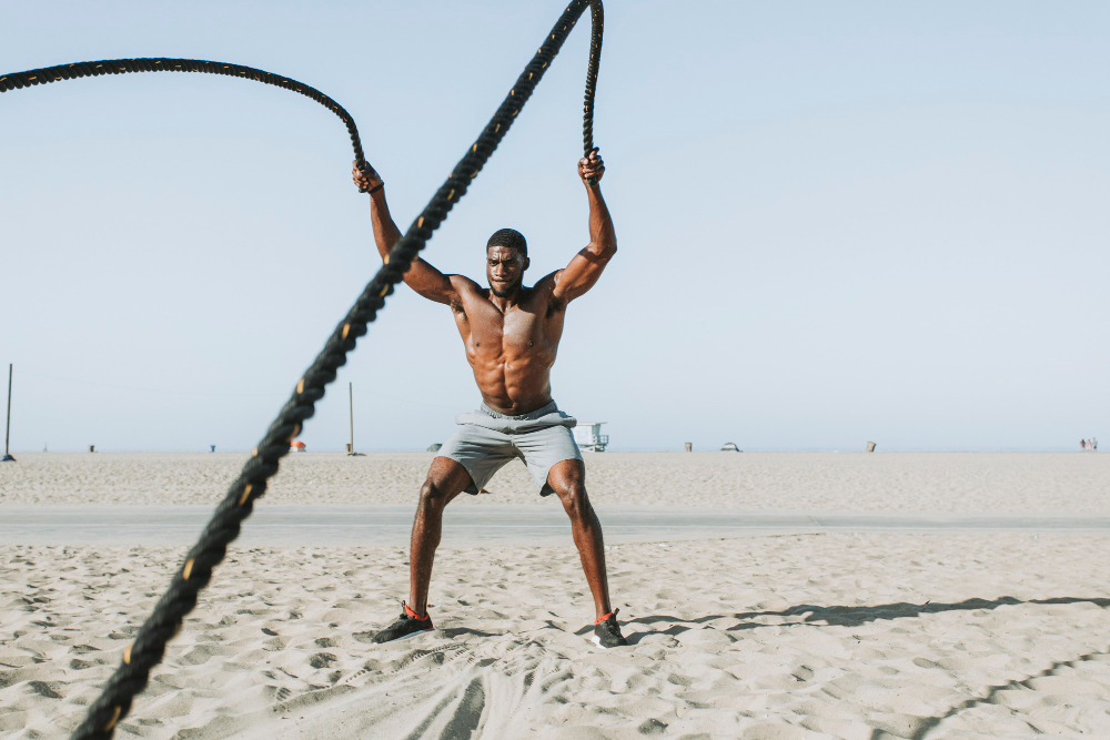 Shirtless man in gray shorts using battle ropes on sandy beach with clear sky.