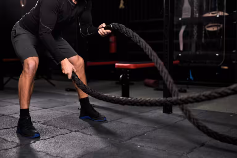 Man in black long-sleeve shirt and shorts using battle rope with blue accent in dark gym with red equipment.