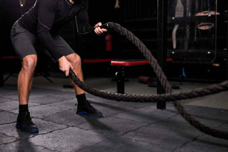 Man in black long-sleeve shirt and shorts using battle rope with blue accent in dark gym with red equipment.