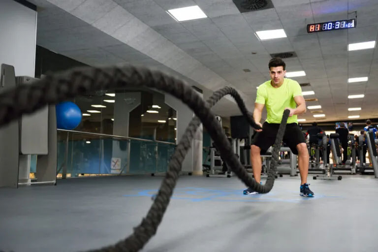 Man in neon yellow shirt using battle ropes in gym with blue exercise ball and stationary bikes visible.