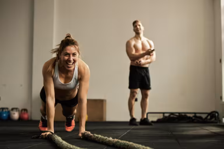 Woman in gray sports bra doing battle rope exercise in gym with man standing behind her near kettlebells.