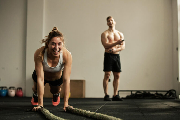 Woman in gray sports bra doing battle rope exercise in gym with man standing behind her near kettlebells.