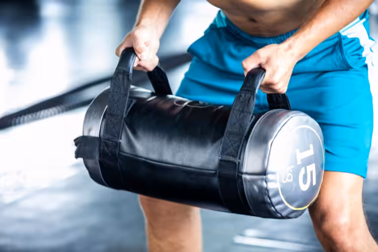 Man wearing blue shorts holding a black, cylindrical 15 kg fitness sandbag by the handles.