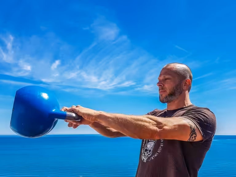 A man with shaved head does a kettlebell swing exercise in front of a bright blue body of water and clear blue sky.