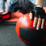 Gloved hand with blue nails resting on a red and black medicine ball during a workout.
