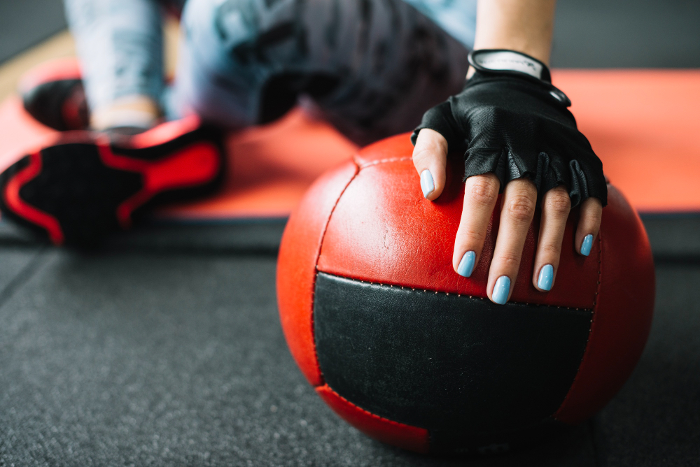 Gloved hand with blue nails resting on a red and black medicine ball during a workout.