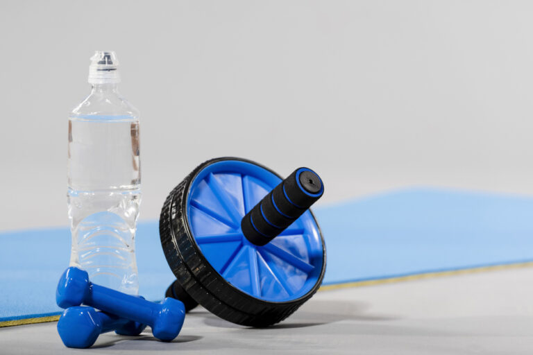 A blue abdominal roller, two small blue dumbbells, and a clear water bottle sit on a light floor by a blue exercise mat.