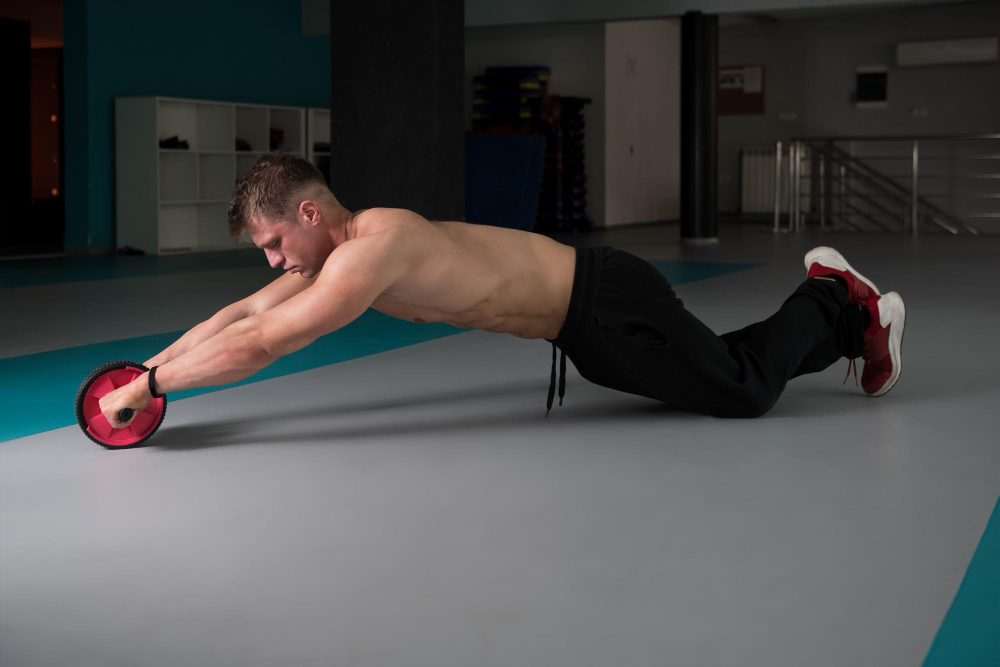 Shirtless man on his knees extending an ab wheel forward in a gym.