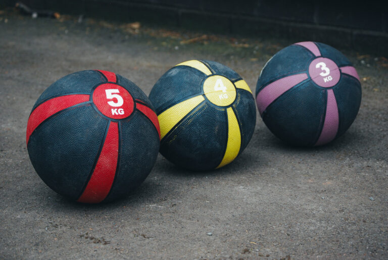 Three medicine balls—red (5kg), yellow (4kg), and purple (3kg)—rest on a dark pavement.
