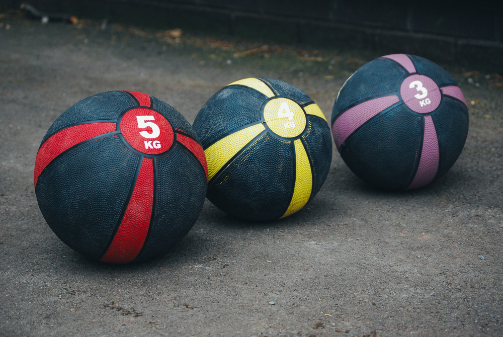 Three medicine balls—red (5kg), yellow (4kg), and purple (3kg)—rest on a dark pavement.