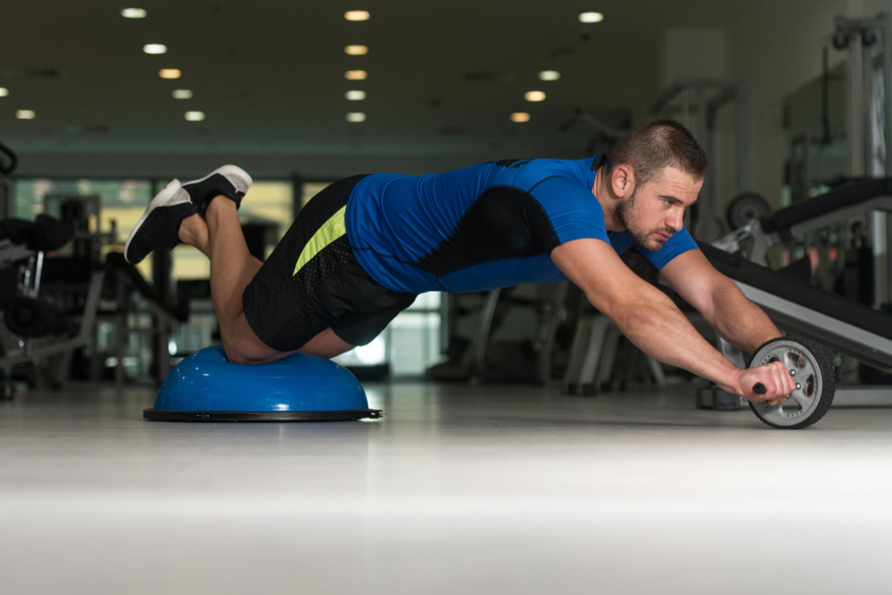 Man balancing on a Bosu ball while extending an ab wheel forward during a gym workout.