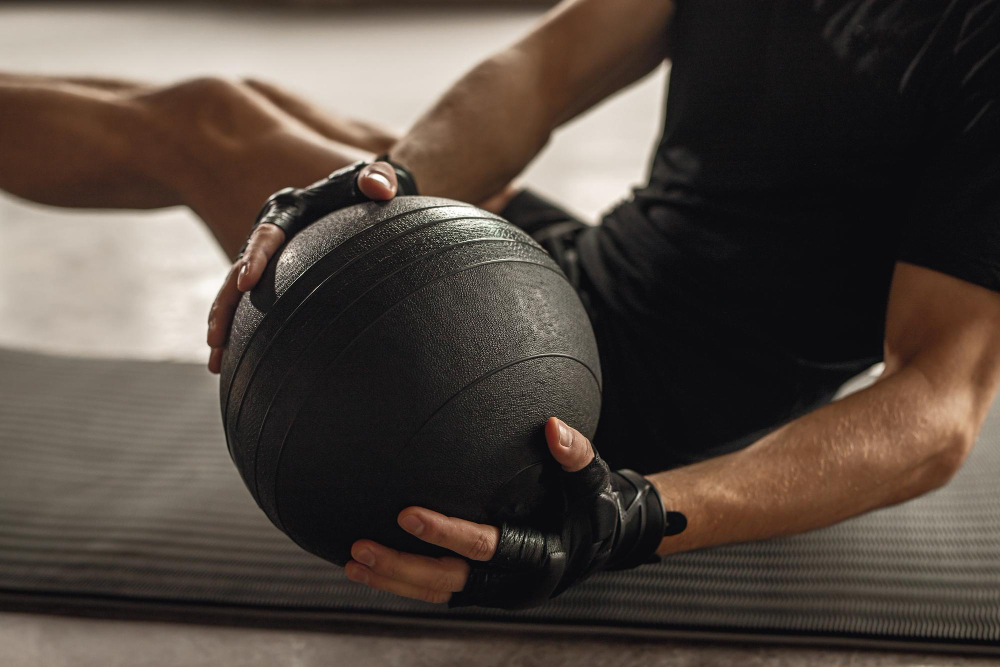 Person seated on a mat holding a black medicine ball with gloved hands during core training.